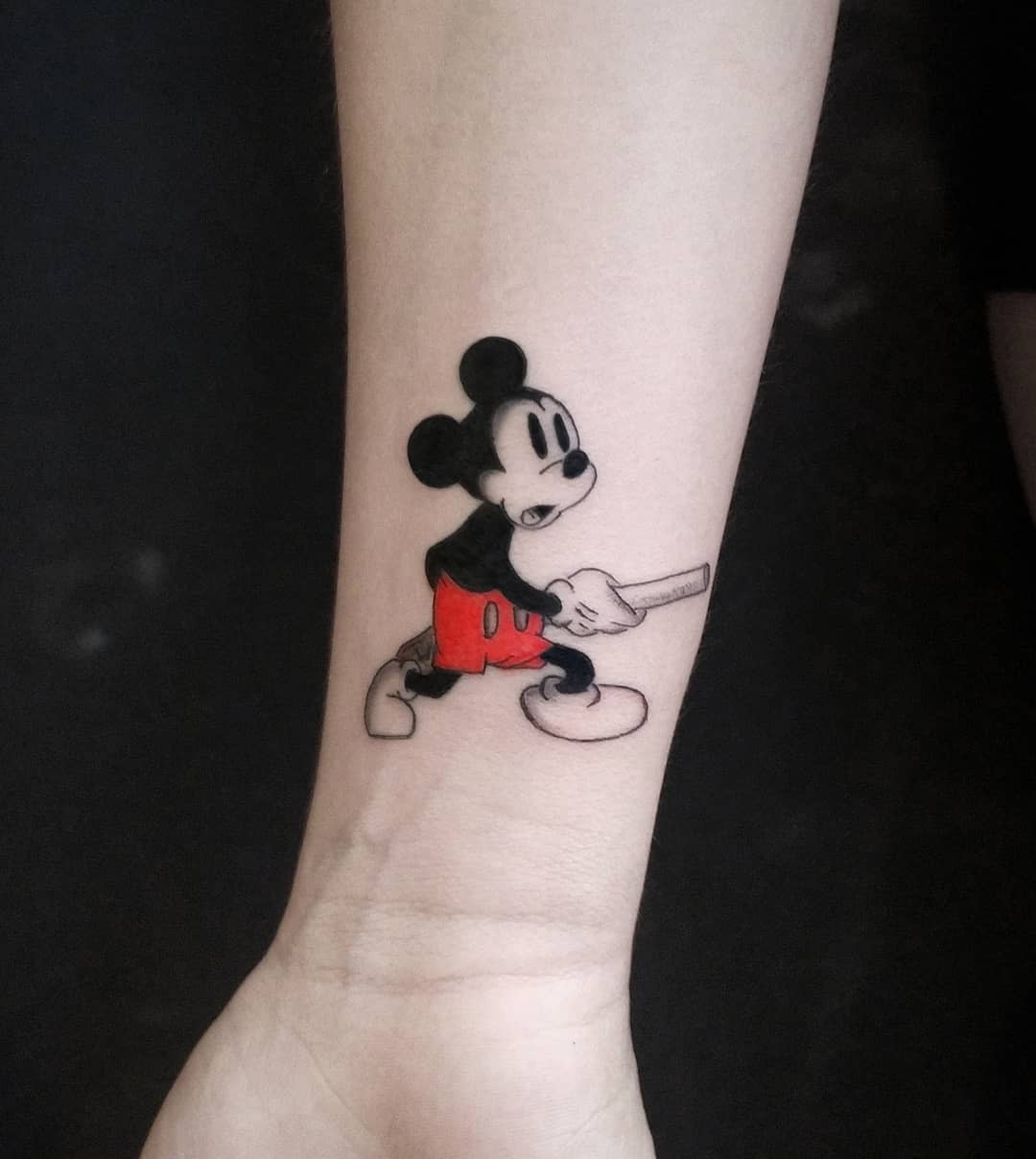 Small tattoo of Mickey Mouse in his classic red shorts, holding a baseball bat, placed on the forearm